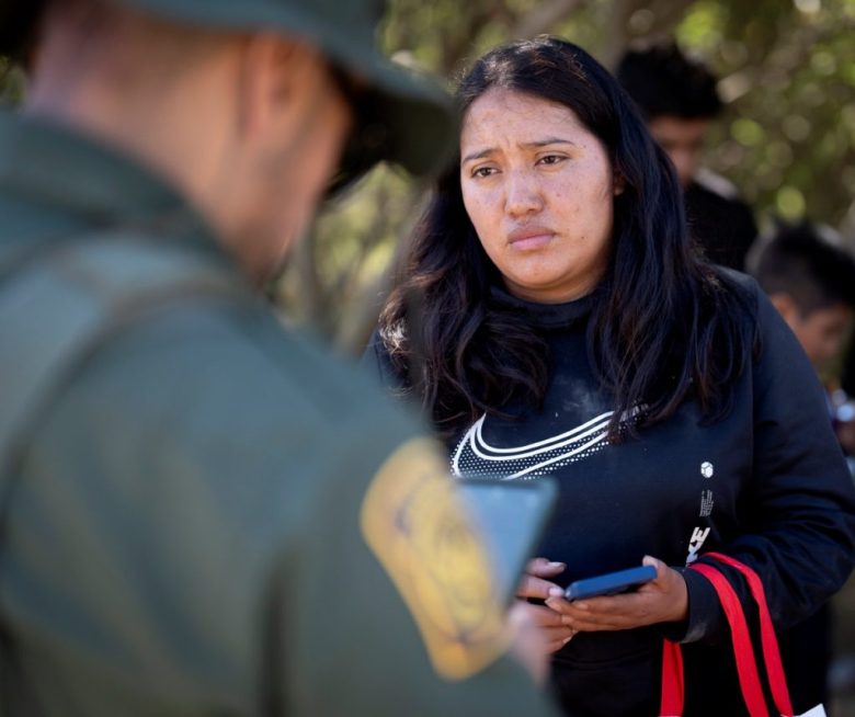 Una migrante de México habla con un agente de la Patrulla Fronteriza antes de ser llevada a una camioneta para ser procesada para el asilo, cerca de Dulzura, California. A la derecha, migrantes que buscan asilo descansan tras cruzar la frontera, en San Diego, California