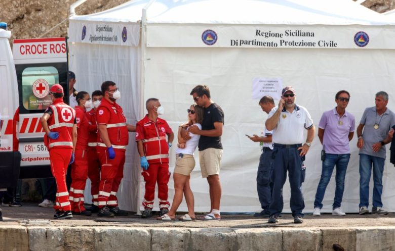 Personas y rescatistas de la Cruz Roja italiana esperan en el muelle mientras los buzos de los bomberos llevan a la orilla el cuerpo de Mike Lynch