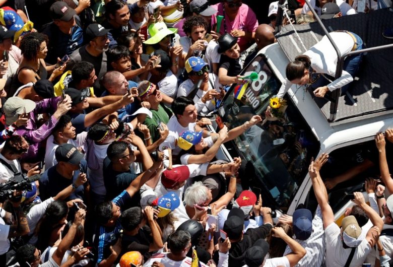 En medio de la multitud, la líder de la oposición venezolana María Corina Machado saluda a seguidores, el sábado, en Caracas. A la izquierda, seguidores de la oposición agitan banderas durante la concentración