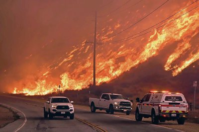 Las llamas devoran colinas cercanas al lago Castaic mientras vehículos transitan sobre la Interestatal 5. A la derecha, una persona moja el techo de su casa con la esperanza de evitar que se destruya