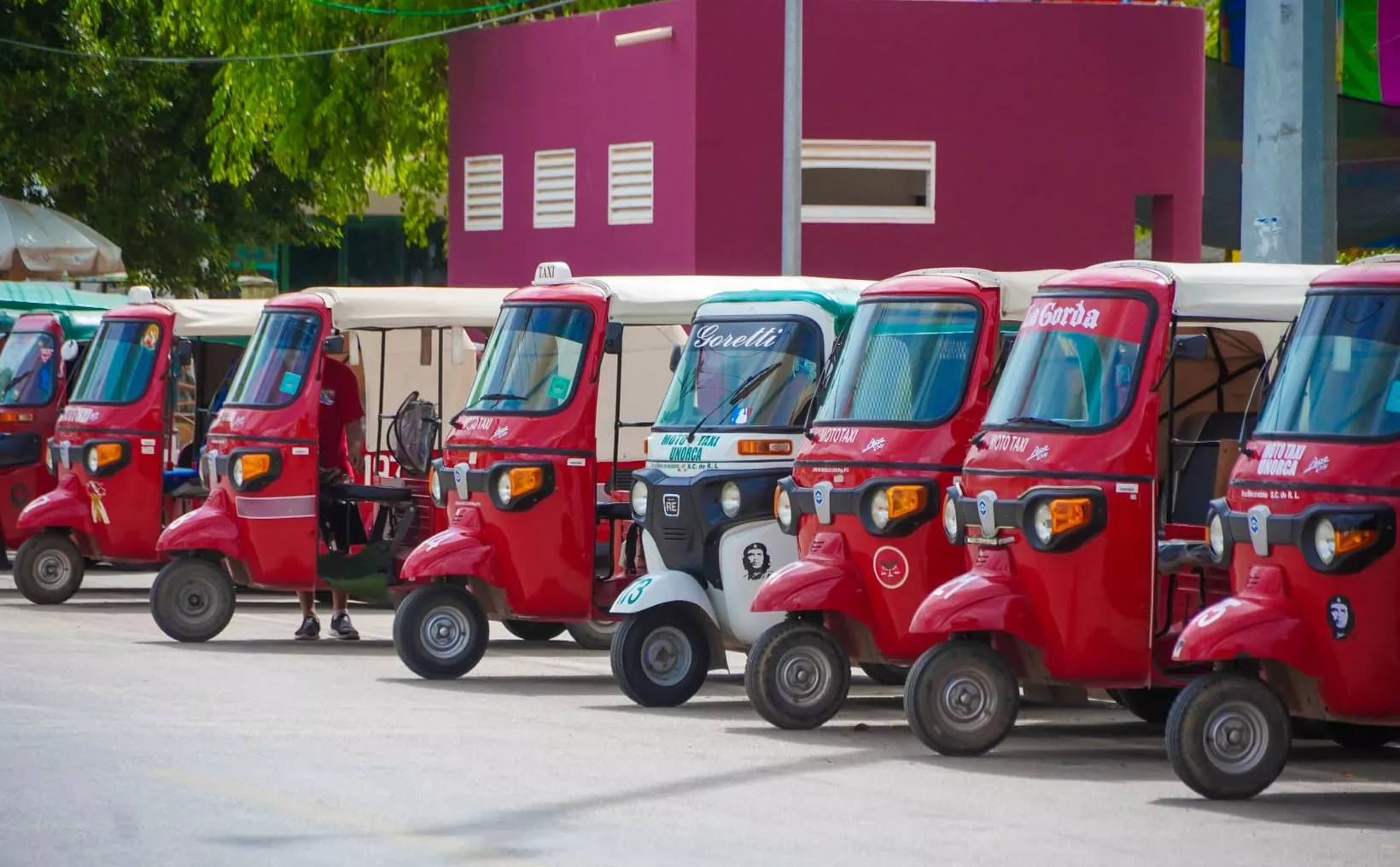 Varios mototaxis estacionados en fila frente a un edificio de color magenta.