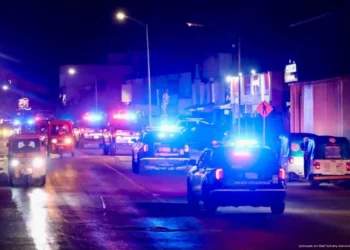 Varias patrullas y vehículos con luces de emergencia circulan de noche por una calle iluminada.