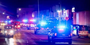 Varias patrullas y vehículos con luces de emergencia circulan de noche por una calle iluminada.