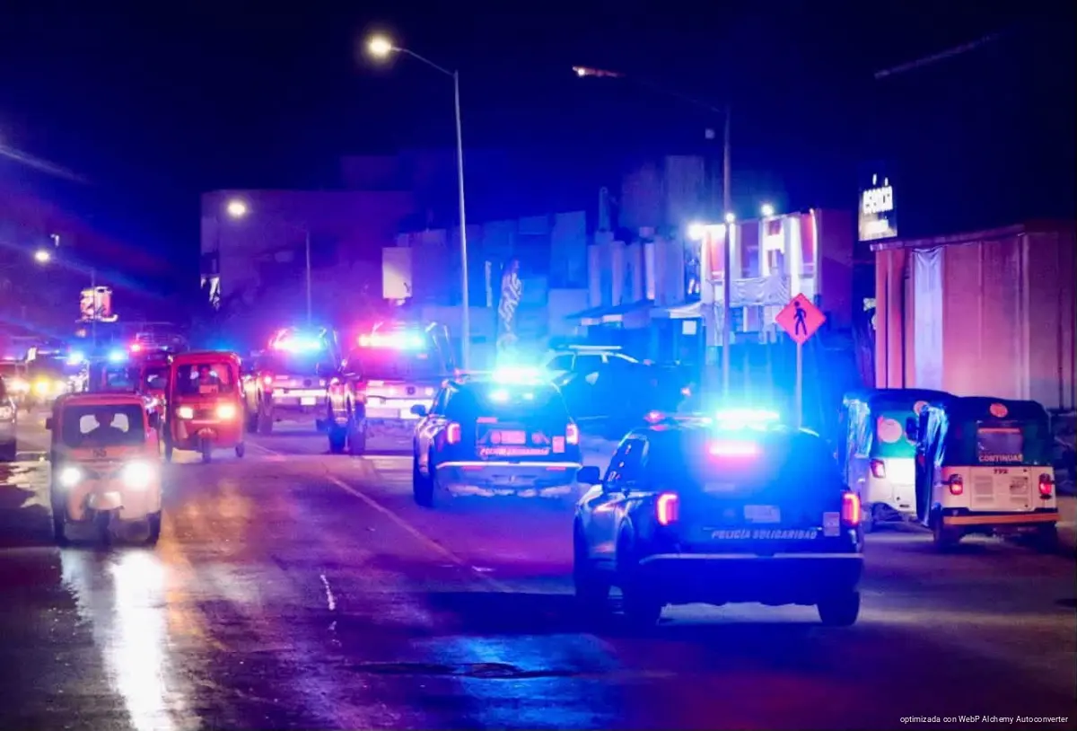 Varias patrullas y vehículos con luces de emergencia circulan de noche por una calle iluminada.