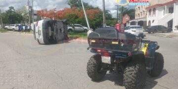 Un vehículo blanco aparece volcado sobre su costado en medio de una calle, mientras una patrulla tipo cuatrimoto está en primer plano y varias personas observan a distancia.
